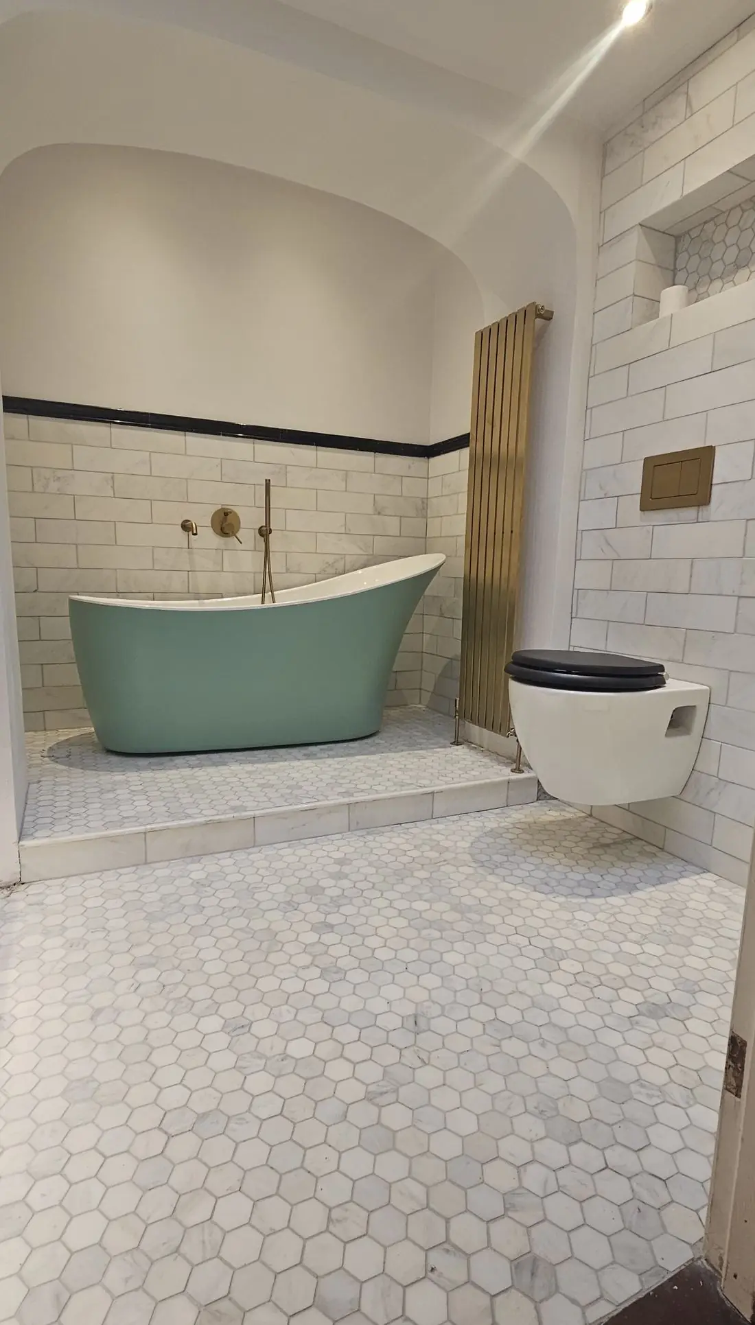 Bathroom with a green freestanding bath in an arched alcove, brass radiator and marble hexagon floor